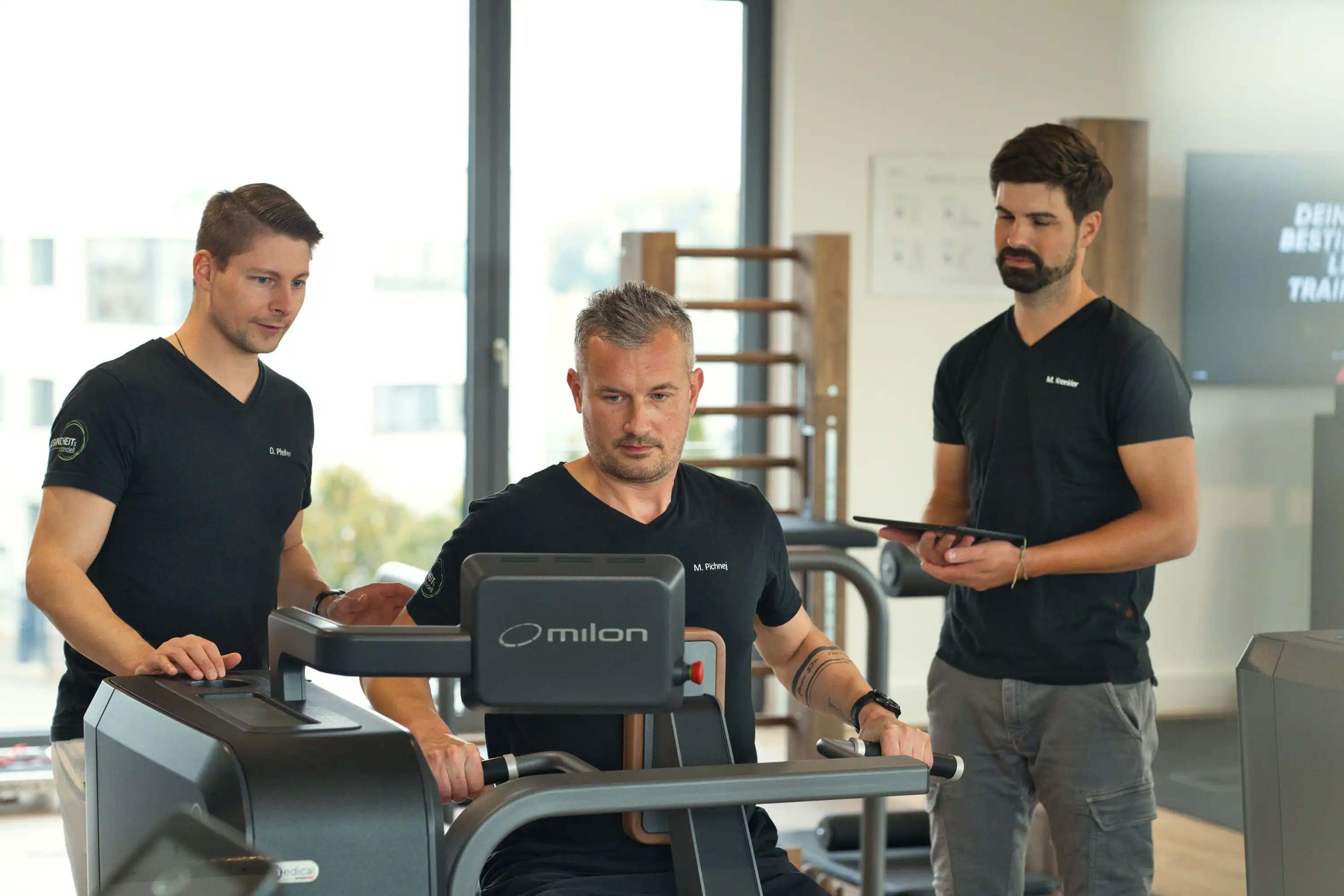 Ganzheitliche Physiotherapie durch enge Zusammenarbeit verschiedener Fachbereiche 2 Physiotherapeuten betreuen einen Patienten an einem Fitnessgerät in einer Physiopraxis in Waiblingen